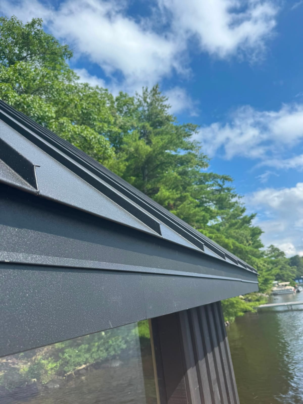 Close-up of a hemmed eave and rake trim detail on a charcoal standing seam metal roof, with a lake and dock in the background