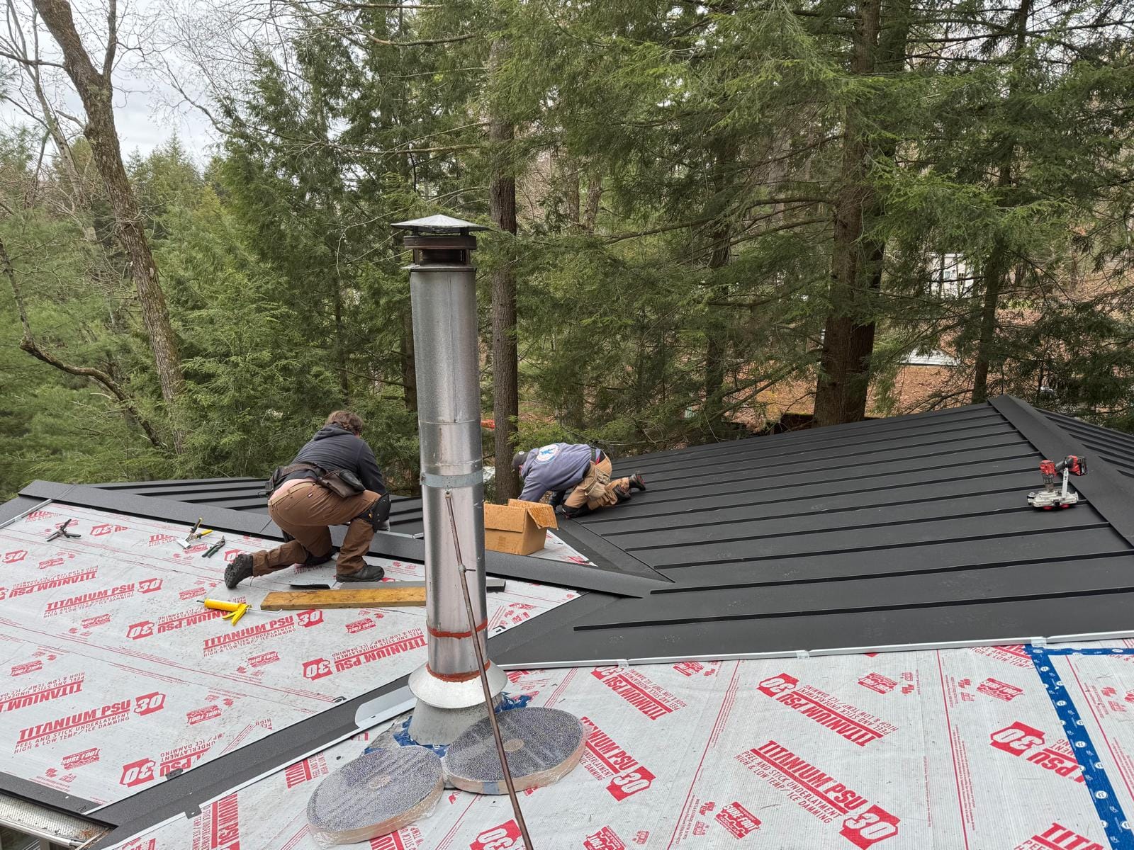 Two CGD Roofing installers fitting charcoal standing seam panels over high-temperature underlayment around a metal chimney flue on a wooded lot