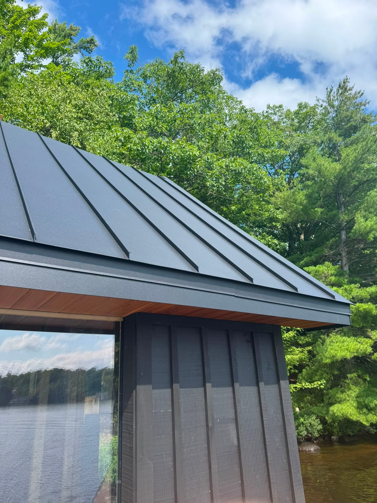 Charcoal standing seam hip roof on a lakeside boathouse with cedar soffit and dark board-and-batten siding, water and forest behind