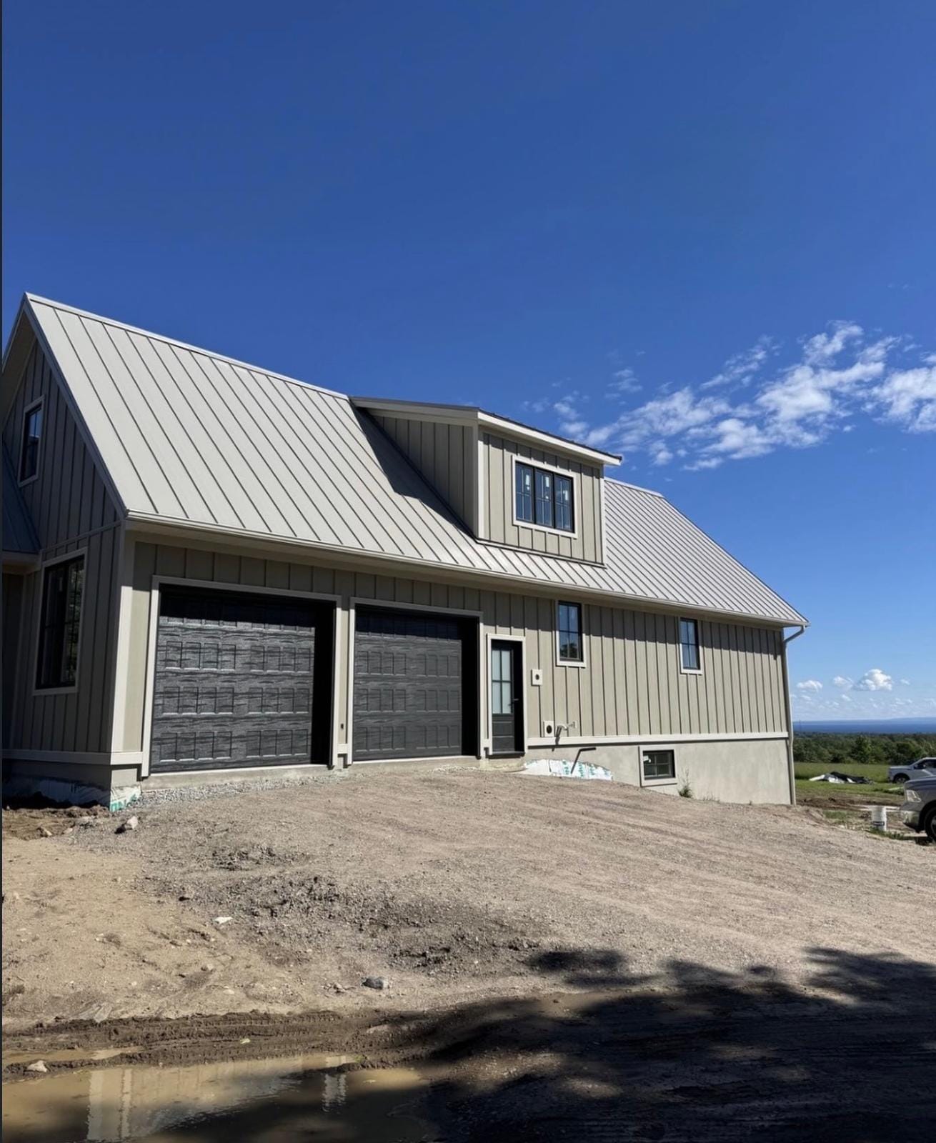 New build home and attached garage with a light grey standing seam metal roof, dormer and board-and-batten siding under a clear blue sky